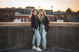Two women showcasing a selection of thrifted women's apparel they purchased at an online thrift store, on a rooftop at sunset.